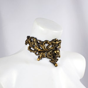 May include: A gold-toned metal choker necklace with an intricate, ornate design. The necklace features a delicate, floral pattern with a pointed, triangular shape.