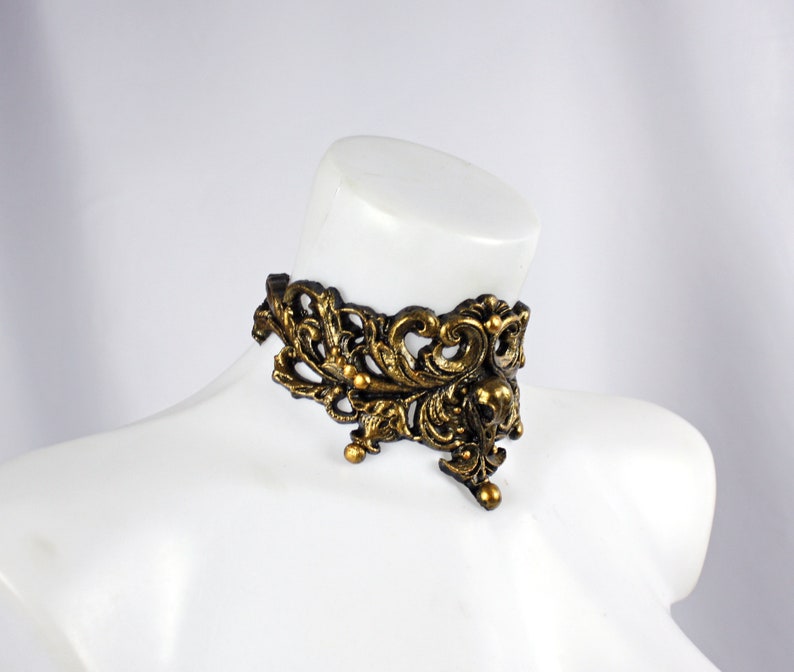 May include: A gold-toned metal choker necklace with an intricate, ornate design. The necklace features a delicate, floral pattern with a pointed, triangular shape.