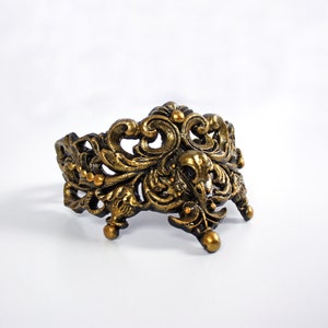 May include: A gold-toned metal ring with an ornate, intricate design. The ring is made of a filigree pattern with a floral motif.