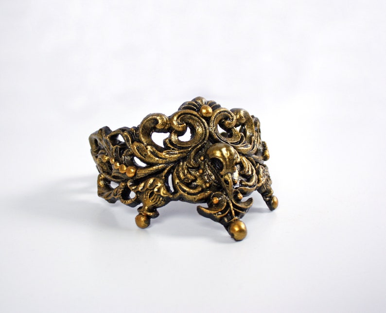 May include: A gold-toned metal ring with an ornate, intricate design. The ring is made of a filigree pattern with a floral motif.