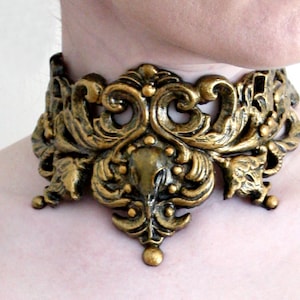 May include: A gold-toned metal choker necklace with an intricate, ornate design. The necklace features a floral pattern with a central motif that resembles a stylized bird.