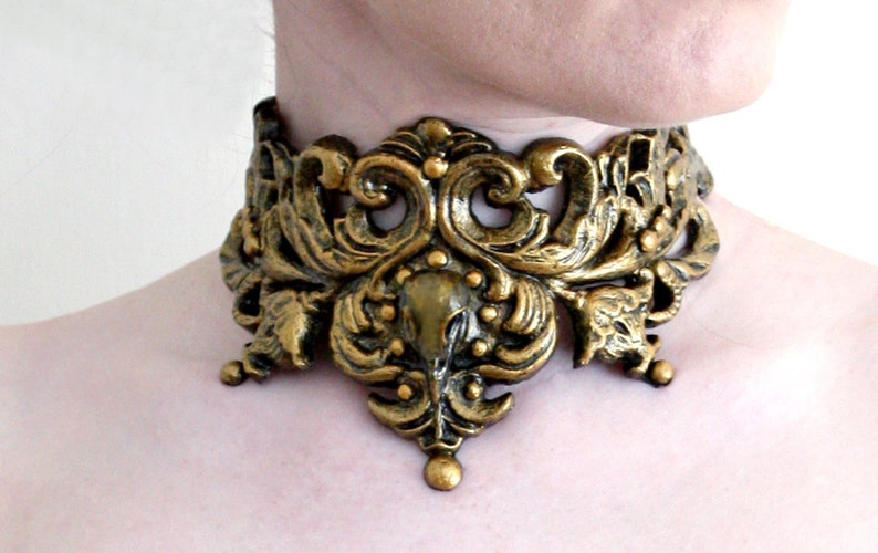 May include: A gold-toned metal choker necklace with an intricate, ornate design. The necklace features a floral pattern with a central motif that resembles a stylized bird.
