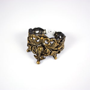 May include: A gold-toned metal bracelet with an intricate, ornate design featuring a floral motif. The bracelet is open on one side and has a clasp on the other.