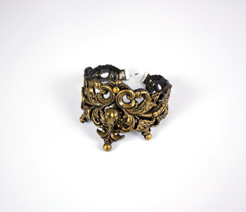 May include: A gold-toned metal bracelet with an intricate, ornate design featuring a floral motif. The bracelet is open on one side and has a clasp on the other.