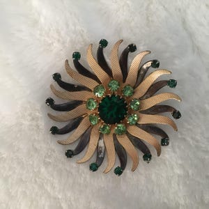 May include: Vintage brooch with a sunburst design. The brooch features a central, large, emerald-green stone surrounded by smaller green stones. The petals are gold and dark brown, creating a striking contrast. A classic piece of jewellery.