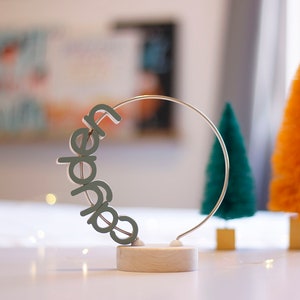 May include: A decorative piece with the word "Jade" in a curved design, mounted on a wooden base. A silver metal ring supports the letters. The background features small, colorful Christmas trees in green and orange, suggesting a holiday theme.
