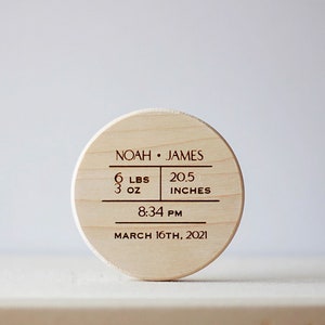 May include: A round wooden keepsake with engraved details. The text reads "NOAH • JAMES" with birth details: 6 lbs 3 oz, 20.5 inches, 8:34 PM, and March 16th, 2021. A personalized baby gift.