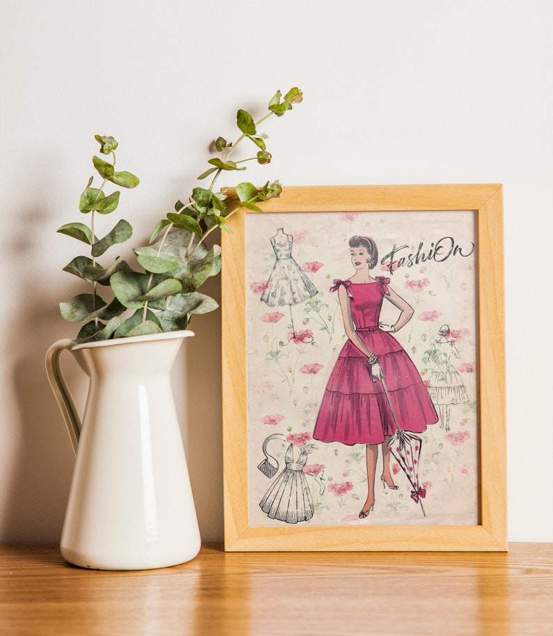 May include: A framed print of a vintage fashion illustration featuring a woman in a pink dress with a white polka dot umbrella. The illustration is surrounded by a floral pattern and the word "Fashion" is written in cursive at the top.