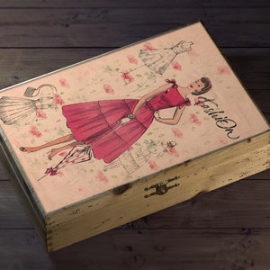 May include: A wooden box with a pink and white floral design featuring a woman in a red dress and the word "Fashion" on the lid.