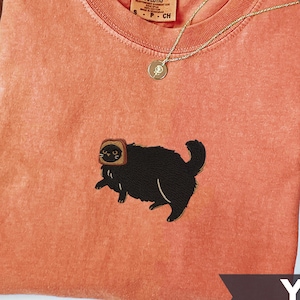 Puede incluir: Una camiseta de color coral con cuello redondo y un gato negro bordado que lleva una rebanada de pan tostado en la cabeza. La camiseta tiene una peque&ntilde;a etiqueta que dice "Comfort Colors". Un collar de oro est&aacute; drapeado sobre la camiseta. La palabra "Yam" est&aacute; impresa en una pancarta.