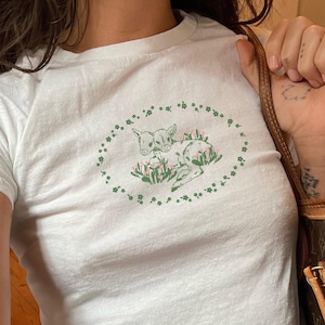 May include: White t-shirt with a green embroidered design of two baby deer in a circle of shamrocks.