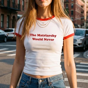 May include: White t-shirt with red trim and the text "The Matriarchy Would Never" in red. The shirt is worn with blue jeans. The background shows a city street with cars and buildings.