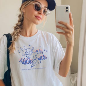 Könnte beinhalten: Ein weißes T-Shirt mit einer blauen und pinkfarbenen Illustration von zwei jungen Rehen mit Sternen und dem Text "There are twice as many stars as sand".