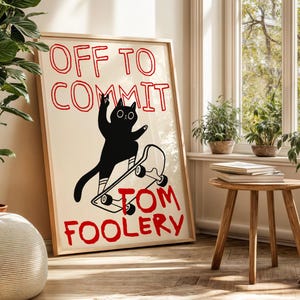 May include: Framed art print featuring a black cat on a skateboard with the text "OFF TO COMMIT TOM FOOLERY" in red. The cat illustration is set against a cream background, framed in natural wood. The artwork is displayed in a well-lit room.
