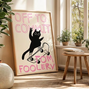May include: A framed art print featuring a black cat skateboarding. The cat is silhouetted against a cream background, with the words "OFF TO COMMIT TOM FOOLERY" in pink. The print is in a light wood frame, leaning against a wall near a window.