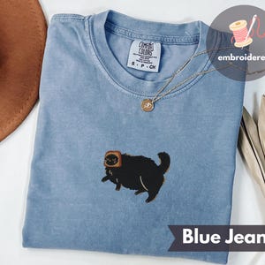 Puede incluir: Camiseta azul vaquero con un dise&ntilde;o bordado de un gato negro con tostadas. La camiseta tiene cuello redondo y mangas cortas. El texto "Blue Jean" est&aacute; en la parte inferior de la imagen. Un collar dorado est&aacute; drapeado sobre la camisa.
