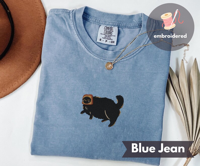 Puede incluir: Camiseta azul vaquero con un dise&ntilde;o bordado de un gato negro con tostadas. La camiseta tiene cuello redondo y mangas cortas. El texto "Blue Jean" est&aacute; en la parte inferior de la imagen. Un collar dorado est&aacute; drapeado sobre la camisa.