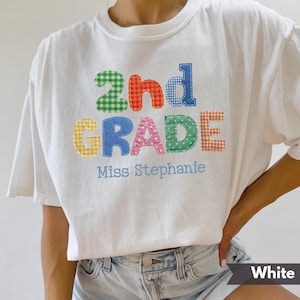以下が含まれることがあります： 「2nd GRADE」と青色の「Miss Stephanie」の文字がカラフルなパッチワークで施された白いTシャツ。文字はギンガムチェックや水玉模様など、様々なパターンで表現されています。カジュアルで着心地の良いTシャツです。
