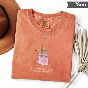 K&ouml;nnte beinhalten: Ein korallenfarbenes Comfort Colors T-Shirt mit Rundhalsausschnitt. Das Shirt zeigt eine Illustration einer Maus mit Krone und rosa Kleid, die einen Zauberstab h&auml;lt. Der Text "I AM THE EXECUTIVE OF THIS DYSFUNCTION" ist darunter gedruckt.