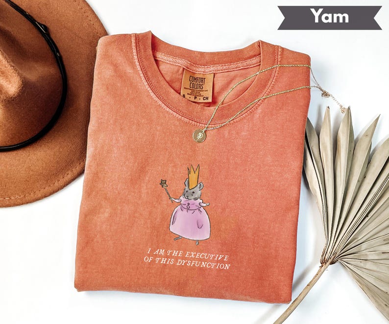 K&ouml;nnte beinhalten: Ein korallenfarbenes Comfort Colors T-Shirt mit Rundhalsausschnitt. Das Shirt zeigt eine Illustration einer Maus mit Krone und rosa Kleid, die einen Zauberstab h&auml;lt. Der Text "I AM THE EXECUTIVE OF THIS DYSFUNCTION" ist darunter gedruckt.