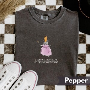 K&ouml;nnte beinhalten: Pfefferfarbenes T-Shirt mit einer skurrilen Illustration einer Maus in einem rosa Kleid und einer Krone, die einen Zauberstab h&auml;lt. Der Text "I AM THE EXECUTIVE OF THIS DYSFUNCTION" ist darunter gedruckt. Wei&szlig;e Turnschuhe sind im Vordergrund.