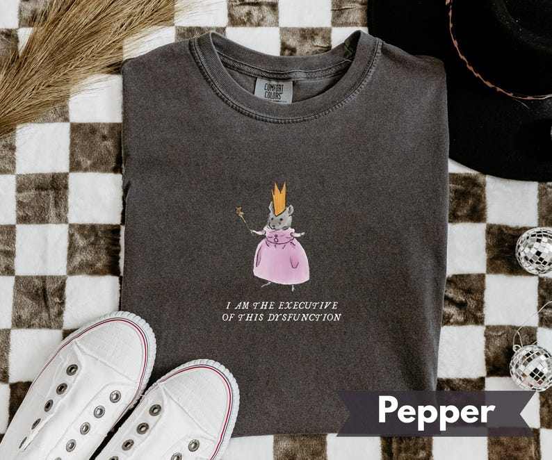 K&ouml;nnte beinhalten: Pfefferfarbenes T-Shirt mit einer skurrilen Illustration einer Maus in einem rosa Kleid und einer Krone, die einen Zauberstab h&auml;lt. Der Text "I AM THE EXECUTIVE OF THIS DYSFUNCTION" ist darunter gedruckt. Wei&szlig;e Turnschuhe sind im Vordergrund.
