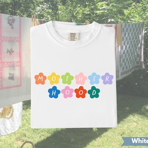 Puede incluir: Una camiseta blanca con la palabra "MOTHERHOOD" escrita en letras con forma de flores de colores. La camiseta está colgada en un tendedero al aire libre, con otras prendas en el fondo.