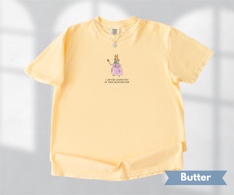 K&ouml;nnte beinhalten: Hellgelbes T-Shirt mit einer Cartoon-Illustration einer Maus als Prinzessin, die einen Zauberstab h&auml;lt. Der Text "I AM THE EXECUTIVE OF THIS DYSFUNCTION" steht darunter. Eine silberne Halskette ist sichtbar.