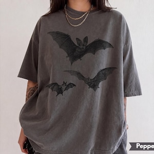 May include: A charcoal gray short-sleeve t-shirt featuring a graphic print of three black bats in flight across the chest. The shirt has a crew neckline and a relaxed, oversized fit. The word Pepper is printed on a small dark banner in the corner.