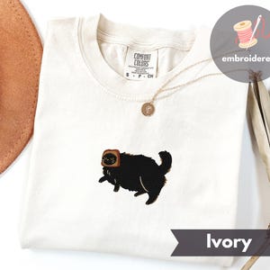Puede incluir: Camiseta de color marfil con un gato negro bordado que lleva una rebanada de pan tostado en la cabeza. La camiseta tiene cuello redondo y mangas cortas. Se ve un collar dorado con un colgante. La camiseta est&aacute; etiquetada como "Comfort Colors".