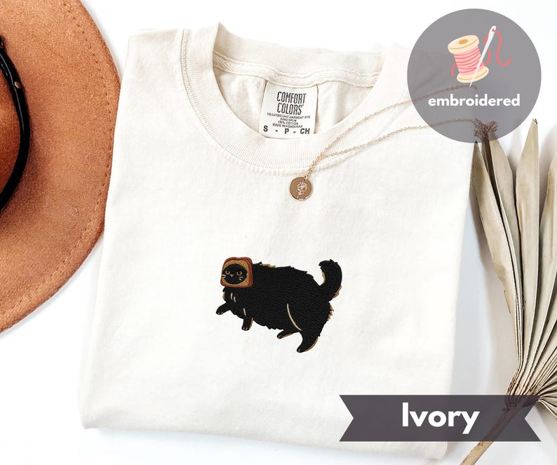 Puede incluir: Camiseta de color marfil con un gato negro bordado que lleva una rebanada de pan tostado en la cabeza. La camiseta tiene cuello redondo y mangas cortas. Se ve un collar dorado con un colgante. La camiseta est&aacute; etiquetada como "Comfort Colors".