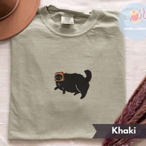 Puede incluir: Camiseta color caqui con un bordado de un gato negro con una rebanada de pan tostado en la cabeza. La camiseta tiene cuello redondo y corte holgado. La palabra "embroidered" est&aacute; en un c&iacute;rculo en la parte superior derecha.