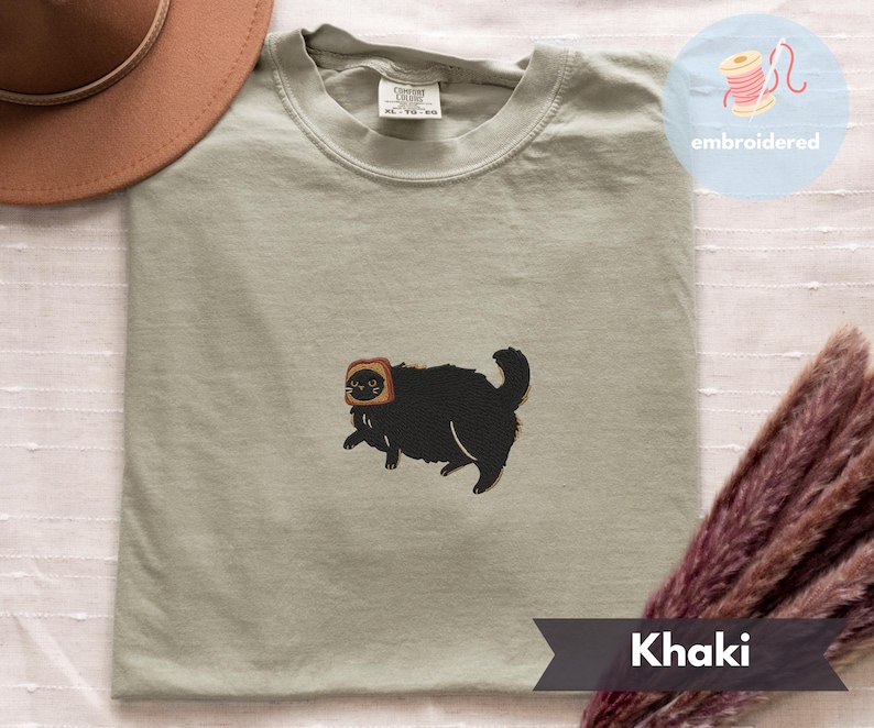 Puede incluir: Camiseta color caqui con un bordado de un gato negro con una rebanada de pan tostado en la cabeza. La camiseta tiene cuello redondo y corte holgado. La palabra "embroidered" est&aacute; en un c&iacute;rculo en la parte superior derecha.