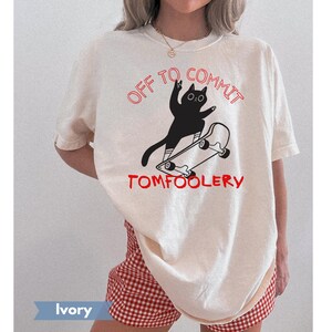 May include: Ivory-colored t-shirt with a black cat graphic riding a skateboard. The text "OFF TO COMMIT TOMFOOLERY" is printed in red above and below the cat illustration. The shirt is paired with red and white checkered shorts.