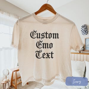 May include: An ivory short-sleeved t-shirt featuring the black text Custom Emo Text in a gothic-style font centered on the chest. The garment is displayed on a wooden hanger and has a crew neckline with a standard fit and hemmed sleeves.