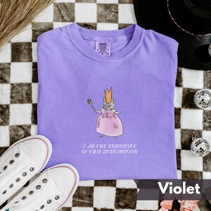K&ouml;nnte beinhalten: Ein violettes T-Shirt mit einer Cartoon-Maus, die eine Krone und ein rosa Kleid tr&auml;gt und einen Zauberstab h&auml;lt. Der Text "I AM THE EXECUTIVE OF THIS DYSFUNCTION" steht darunter. Wei&szlig;e Turnschuhe befinden sich unten links.