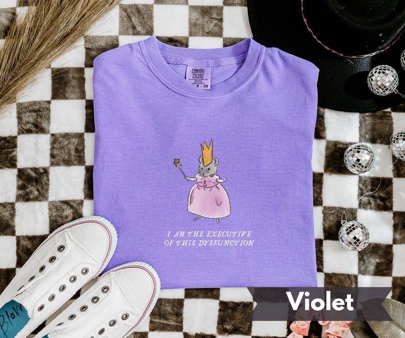 K&ouml;nnte beinhalten: Ein violettes T-Shirt mit einer Cartoon-Maus, die eine Krone und ein rosa Kleid tr&auml;gt und einen Zauberstab h&auml;lt. Der Text "I AM THE EXECUTIVE OF THIS DYSFUNCTION" steht darunter. Wei&szlig;e Turnschuhe befinden sich unten links.