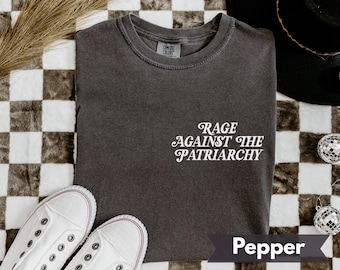 May include: A dark gray t-shirt with the text "RAGE AGAINST THE PATRIARCHY" in white script. The shirt is folded on a checkered surface, with white sneakers and a black hat visible. The shirt color is "Pepper".