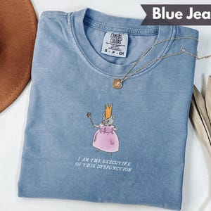 K&ouml;nnte beinhalten: Blaues Jeans-T-Shirt mit einer Grafik einer Maus mit Krone und rosa Kleid, die einen Zauberstab h&auml;lt. Der Text "I AM THE EXECUTIVE OF THIS DYSFUNCTION" ist darunter gedruckt. Eine goldene Halskette liegt auf dem Shirt.