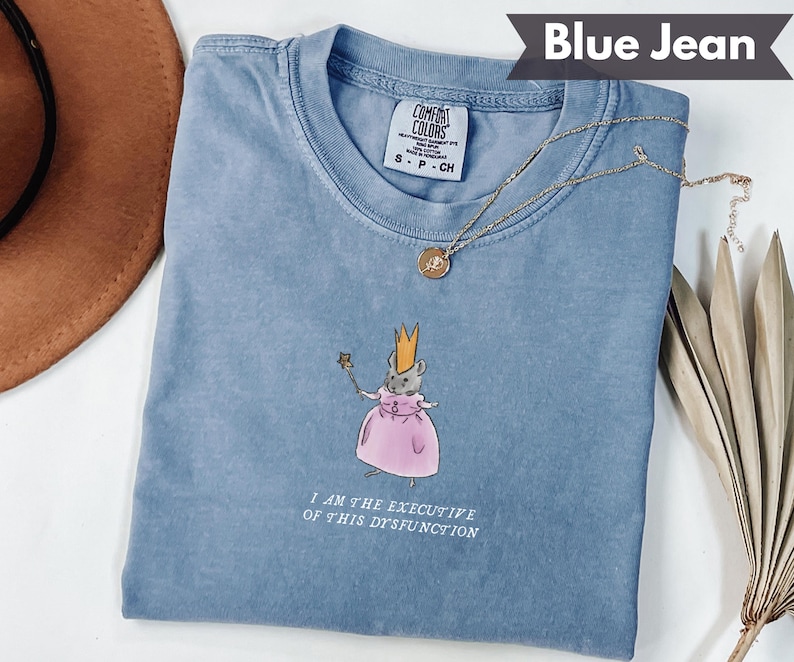 K&ouml;nnte beinhalten: Blaues Jeans-T-Shirt mit einer Grafik einer Maus mit Krone und rosa Kleid, die einen Zauberstab h&auml;lt. Der Text "I AM THE EXECUTIVE OF THIS DYSFUNCTION" ist darunter gedruckt. Eine goldene Halskette liegt auf dem Shirt.