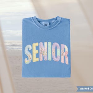 May include: A blue, short-sleeved shirt with the word "SENIOR" in colorful, patchwork-style letters. The letters have floral and pastel patterns. The shirt is made of a soft material and is perfect for graduation.