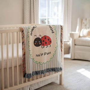 May include: A woven throw blanket featuring a red ladybug with black spots, a green scalloped border, and blue vertical stripes. The text reads "mommy's lovebug" and "sadie grace" in black script. The blanket has fringed edges on the sides.
