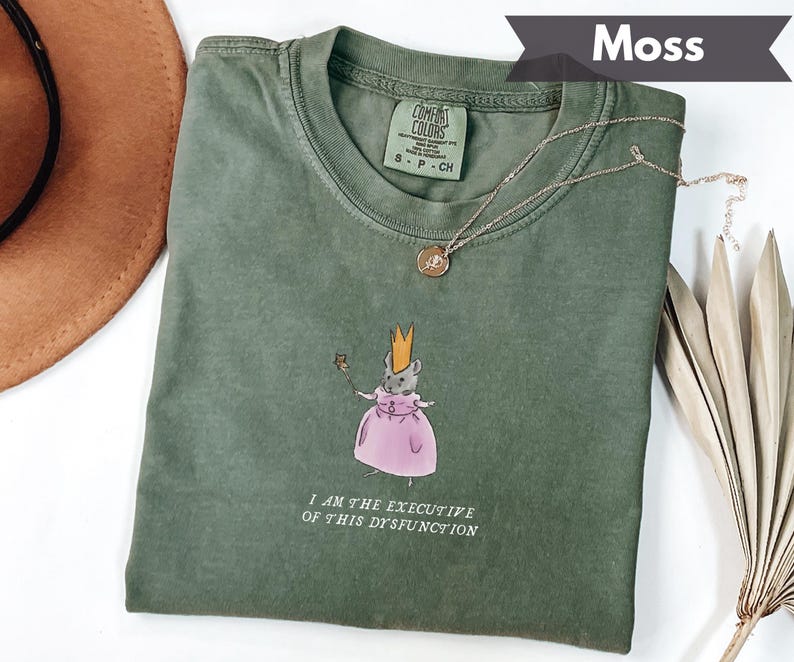 May include: Moss green t-shirt with a graphic of a mouse wearing a crown and pink dress, holding a wand. The text "I AM THE EXECUTIVE OF THIS DYSFUNCTION" is printed below the graphic. A gold necklace is draped across the shirt.