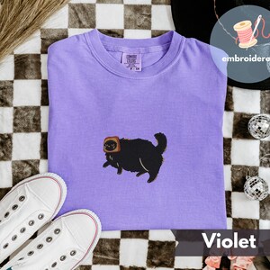 Puede incluir: Camiseta violeta con un bordado de un gato negro con una tostada en la cabeza. La camiseta est&aacute; doblada sobre una superficie a cuadros, con zapatillas blancas y bolas de discoteca. La palabra "embroidered" est&aacute; escrita en una pegatina redonda.