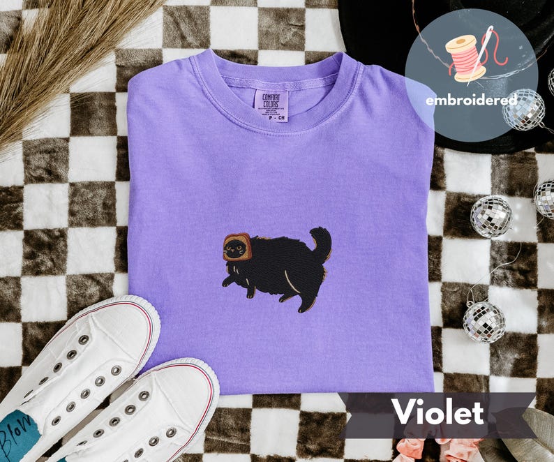 Puede incluir: Camiseta violeta con un bordado de un gato negro con una tostada en la cabeza. La camiseta est&aacute; doblada sobre una superficie a cuadros, con zapatillas blancas y bolas de discoteca. La palabra "embroidered" est&aacute; escrita en una pegatina redonda.