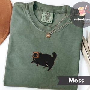 Puede incluir: Camiseta Comfort Colors verde musgo con un gato negro bordado con tostadas. La camiseta tiene cuello redondo y mangas cortas. Se ve un collar plateado con un colgante. La palabra "Moss" est&aacute; impresa en una pancarta.