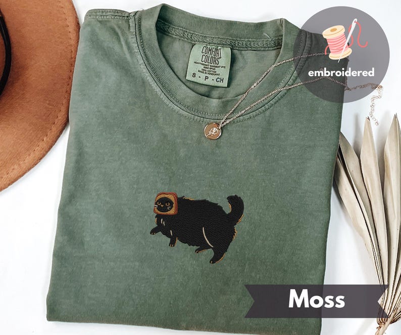 Puede incluir: Camiseta Comfort Colors verde musgo con un gato negro bordado con tostadas. La camiseta tiene cuello redondo y mangas cortas. Se ve un collar plateado con un colgante. La palabra "Moss" est&aacute; impresa en una pancarta.