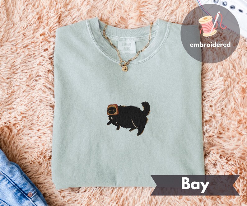 Puede incluir: Sudadera de cuello redondo verde claro con un gato negro bordado con una rebanada de pan tostado en la cabeza. Una cadena dorada cuelga sobre la camisa. La palabra "Bay" est&aacute; impresa en una pancarta en la parte inferior.