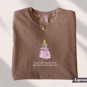 K&ouml;nnte beinhalten: Espressofarbenes Sweatshirt mit einer skurrilen Illustration einer Maus mit Krone und rosa Kleid, die einen Zauberstab h&auml;lt. Der Text "I AM THE EXECUTIVE OF THIS DYSFUNCTION" ist darunter gedruckt. Eine goldene Halskette liegt auf dem Shirt.