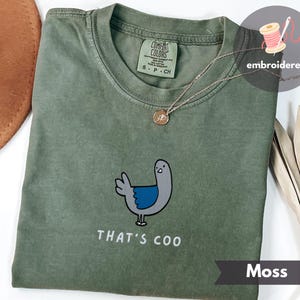 Könnte beinhalten: Moosgrünes T-Shirt mit einem gestickten Cartoon-Taubendesign. Die Taube ist grau mit blauen Akzenten und dem Text "THAT'S COO" in Weiß. Eine goldene Halskette ist sichtbar. Das Shirt ist mit "Comfort Colors" gekennzeichnet.